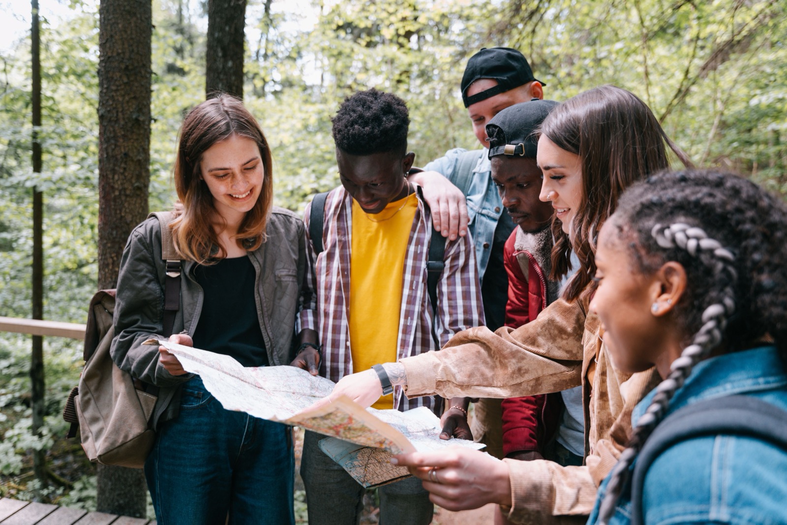 Six friends standing together on a forest trail, gathered around two open paper maps as one of them points out a route.
